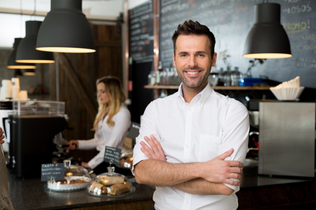 small business owner standing with crossed arms with employee in background preparing coffee
