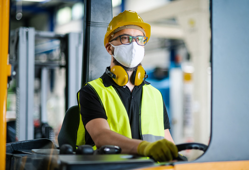 A forklift driver in an industrial factory wearing a hard hat, a mask, noise-suppression headphones, gloves, and a high-visibility vest